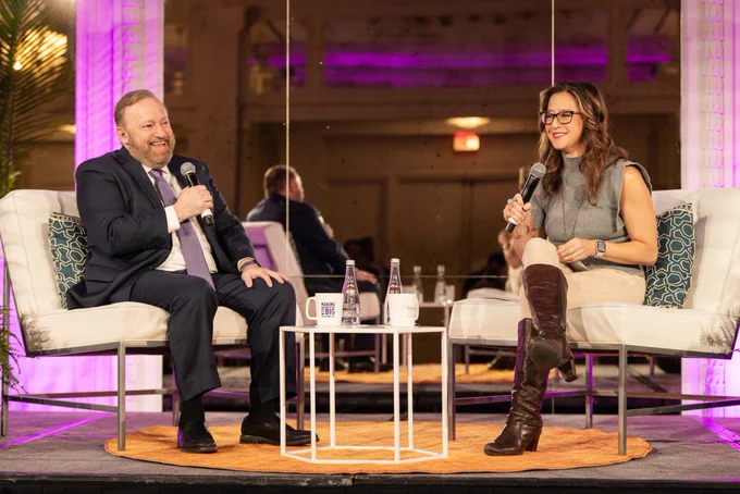 Two people smile and converse on a stage.