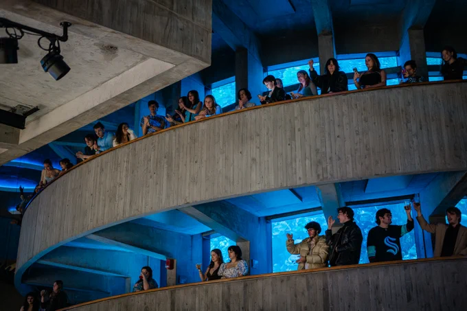 People cheer and clap on a winding balcony that wraps a large fish tank.