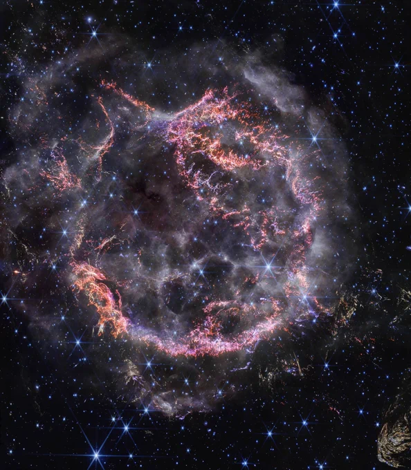  A roughly circular cloud of gas and dust with complex structure. The inner shell is made of bright pink and orange filaments studded with clumps and knots that look like tiny pieces of shattered glass. Around the exterior of the inner shell, there are curtains of wispy gas that look like campfire smoke. Around and within the nebula, various stars are seen as points of blue and white light. Outside the nebula, there are also clumps of dust, coloured yellow in the image.