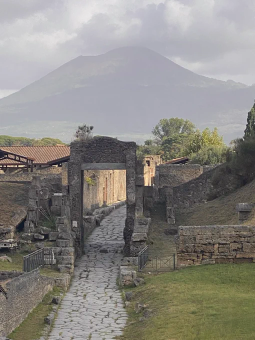 A view up the Roman road that runs under the Nocera Gate first thing in the morning and a wall in the city is spotlit in morning sun while Vesuvius behind looks moody as clouds gather above its summit