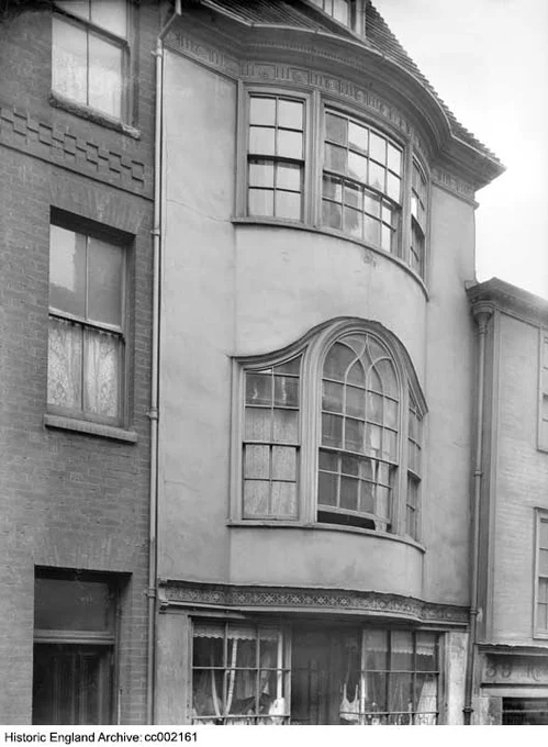 A black and white photograph of the upper floors of a three storey, one bay wide shop. At the bottom of the photograph, the upper part of a multi-paned shop window is visible. The upper two floors above have three-light bow windows. The front of the building is faced in a smooth render. 