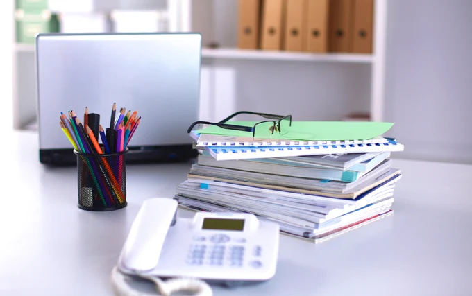 Image of desk, books, glasses, laptop, pot of pens and phone