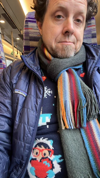 Peter sitting in an Elizabeth line train in a colorful scarf and Christmas jumper with a silly grimace on his face.