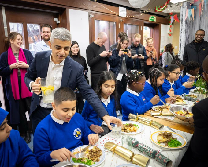 Mayor of London helps serve drinks to primary school children who are sitting eating their lunch at dinner tables surrounded by Christmas crackers in a decorated hall.

