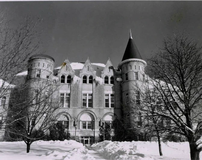 A black and white photo of a building. Snow and icicles can be seen on the rooftop of the building. Snow and dead trees are in front of the building.  