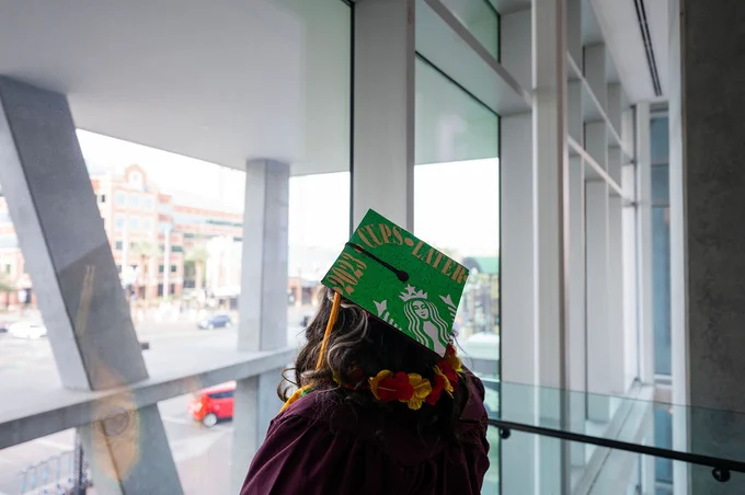 A graduating student, wearing a grad cap reading “2023 cups later” and adorned with the Starbucks logo, looks out the window toward ASU.