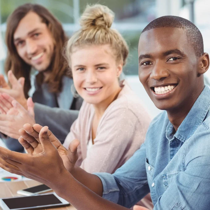 Three people sit in a row at a desk, looking into the camera, smiling and clapping