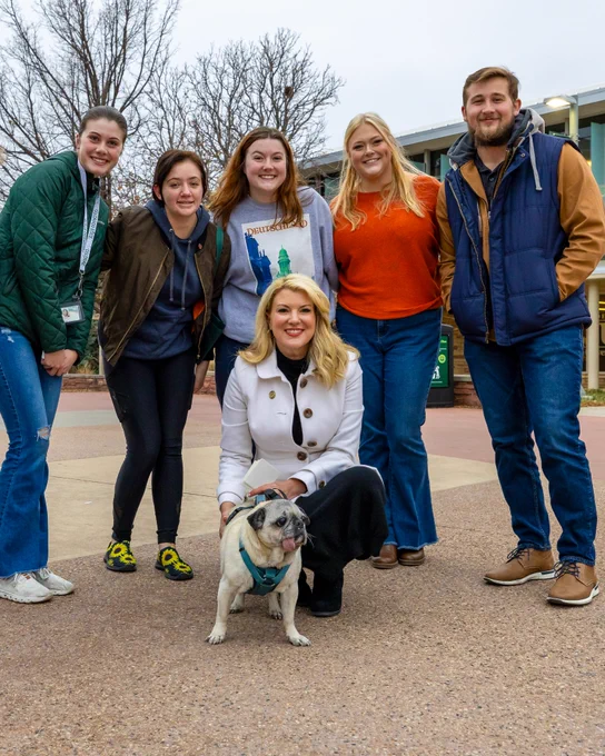 President Amy Parsons knelt down beside her pug, as five students stand behind her posing for a picture on the Plaza at C S U.