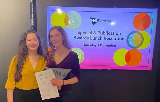 Dr Leanne Jones and Rosanna Roskilly standing together and smiling.  Rosanna is holding her IoM3 award and certificate.  Behind them is an LED screen displaying the IoM3 logo and the words 'Special & Publication Awards Lunch Reception Thursday 7 December' with coloured circles decorating the border of the screen.