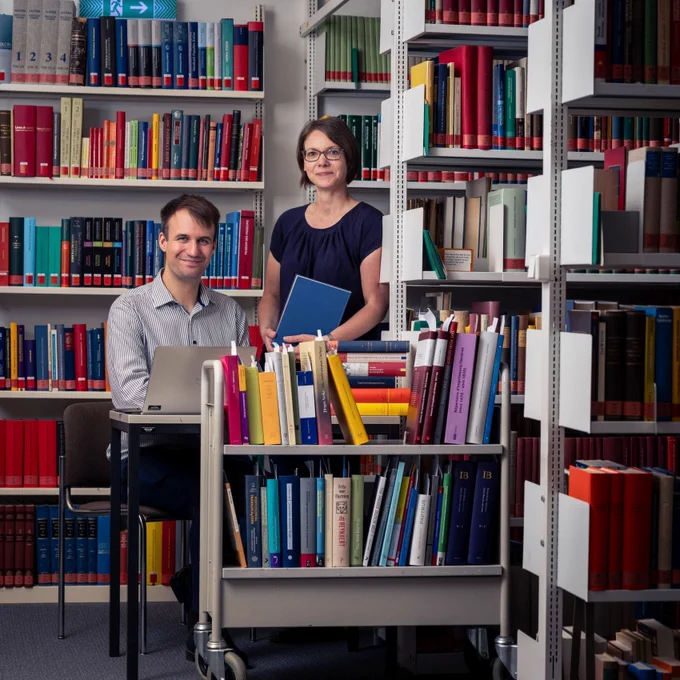Die Abbildung zeigt Henning Ohst und Sandra Simon, umringt von Bücherregalen. Henning Ohst sitzt an einem Tisch und vor ihm steht ein aufgeklappter Laptop. Sandra Simon steht daneben und hält ein Buch in der Hand. Beide blicken lächelnd in die Kamera. Vor den beiden steht ein voller Bücherwagen mit Büchern aus denen an der Oberseite weiße Leihzettel hervorschauen.