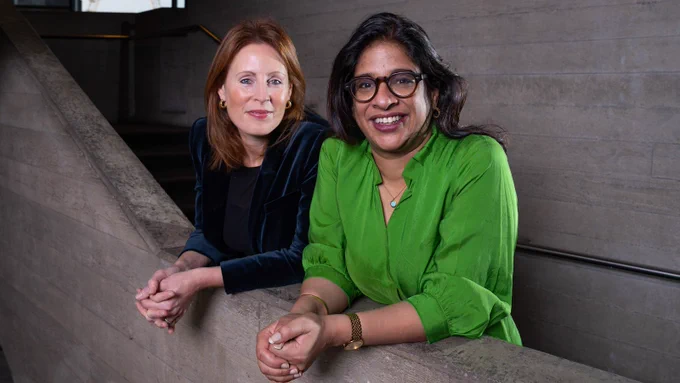 Kate Varah and Indhu Rubasingham stand side by side, leaning on a concreate staircase bannister within the National Theatre, smiling at the Camera.