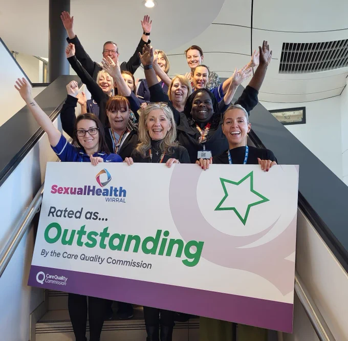 NHS staff stood on a stairwell holding banner: Sexual Health Wirral rated as Outstanding by the Care Quality Commission