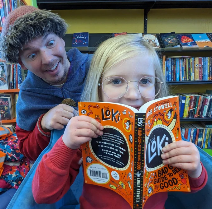 A young girl holding open a book with a man dressed in Viking attire stood behind her.