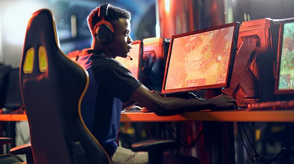 A young person wearing headphones with an attached microphone sits on a high backed chair holding a keyboard mouse and looking at a computer screen showing video game footage