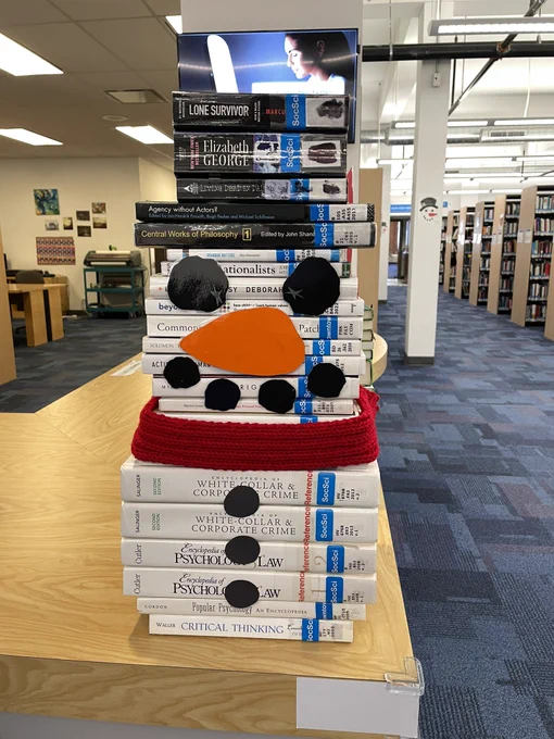 Books piled on top of one another in the shape of a snowman with a black top hat. Black circles of construction paper are placed on the books to make eyes, a mouth and three buttons with an orange paper carrot nose. It also has a red scarf.