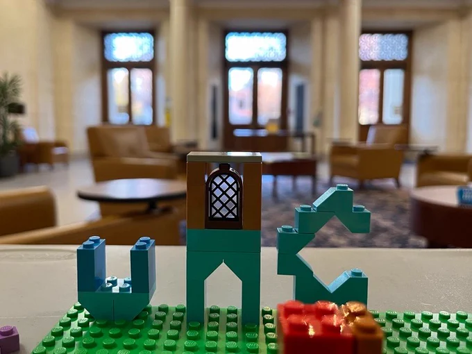 In the foreground, an assemblage of teal, green, orange, and purple logos. The teal pieces are shaped into the initials UNC. A Lego window piece is placed on top of the N in UNC. In the background, a blurred view of the Wilson Library Lobby.