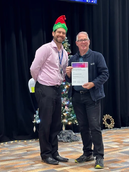 Two people standing at the front of a room with one holding an award certificate