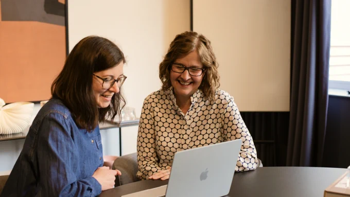 Two women sitting side by side are looking down at a laptop screen. They both have brown hair, glasses and are smiling.