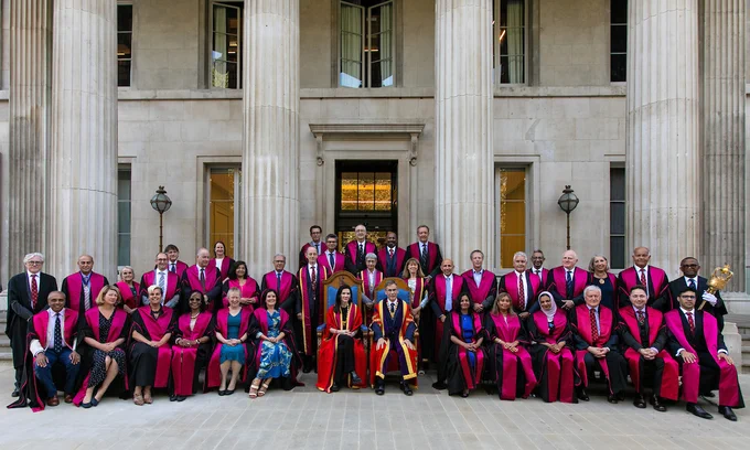 The RCS England Council sit for an official photograph outside of the RCS England building. They all wear black ceremonial robes which are lined in bright pink. 