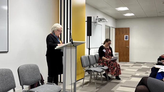 Mrs Kay Martin stands at a podium in a black skirt suit and boots. She reads from the podium. In the background Laurence Totelin sits on a chair looking at the print programme. She wears a red flowery dress.