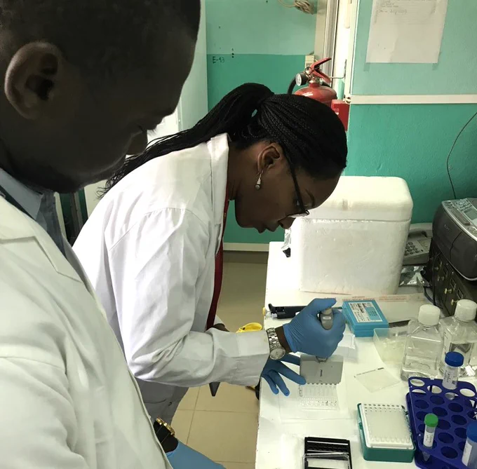 Dr Kone is pipetting samples in a lab with a jade blue painted wall. She is wearing blue gloves and a white lab coat. There are various laboratory equipment and reagents on the counter top. A colleague, also in a white lab coat, is watching her actions.