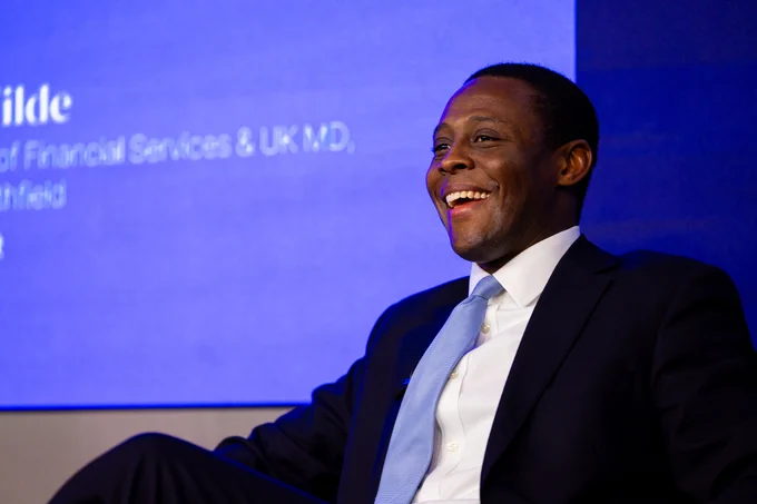 City Minister Bim Afolami speaks at the London Stock Exchange as part of Investor Summit 2023. He is smiling towards the audience with a blue screen behind him.