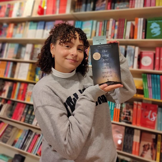 Our Isabelle, standing in front of our The Hague store's General Fiction section and holding up a copy of The Long Way to a Small, Angry Planet by Becky Chambers. She is smiling at the camera and holding the book up besides her head with two hands.