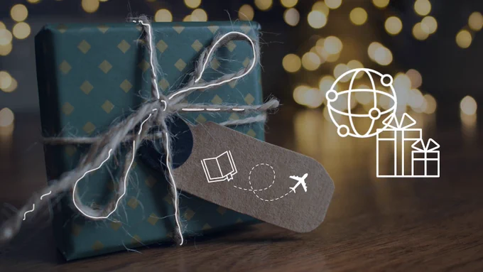 A present in front of lights showing in the background. The present has a paper tag showing the illustrations of a book and an airplane.