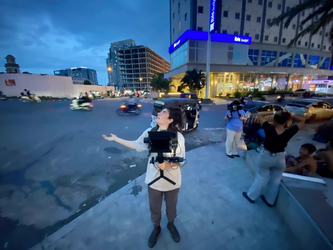 It's twilight in a city and a woman with a camera stands looking to the sky, one arm out checking for rain