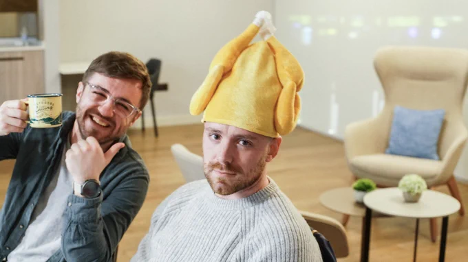 Two men smiling at the camera, one holding up a mug and one with a turkey hat on.