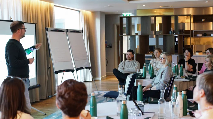 Man stood at the front of a room talking. People sat around tables.