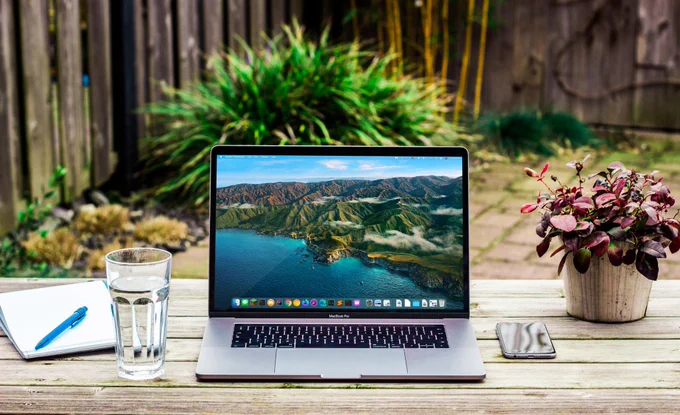 A laptop on an outdoor bench, surrounded by green plants. 