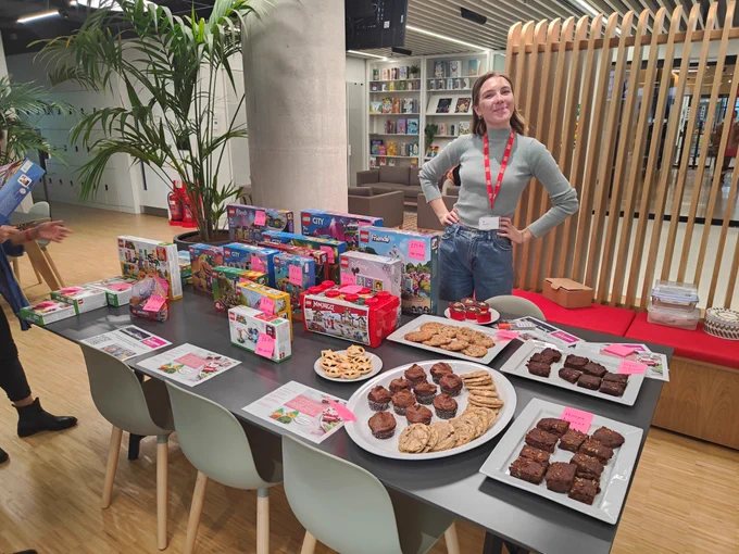 A woman stands behind a stall selling cakes, biscuits and boxed LEGO sets in an office lobby. 