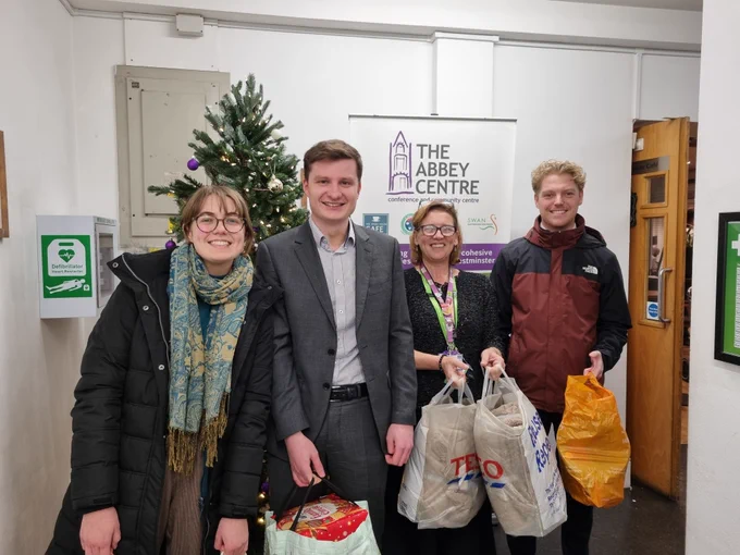 Four people smiling and standing in front of a Christmas tree holding bags full of gifts.