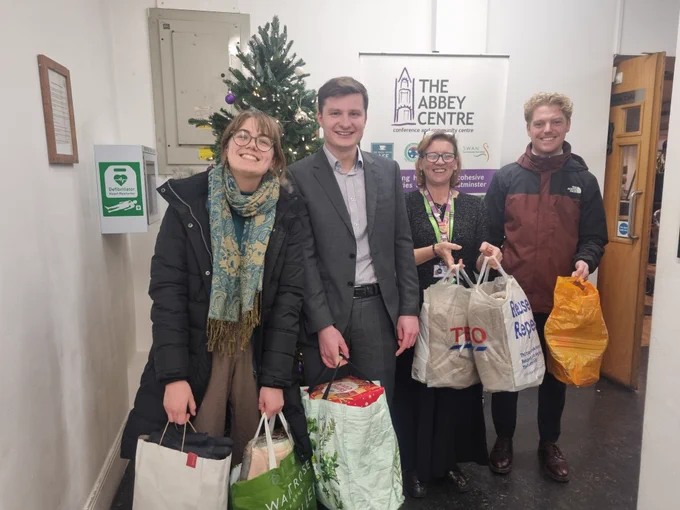 Four people smiling and standing in front of a Christmas tree holding bags full of gifts.