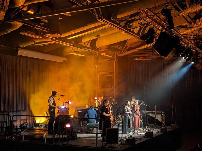 The Unthanks onstage at Sheffield's Octagon Centre. Orange uplighting onto air conditioning pipes, white spotlights on the singers.