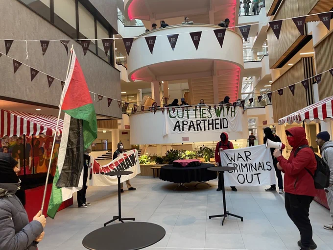 Students in the ground floor of the Owen building atrium with Palestine flags and banners that say ‘¡NO PASARÀN!’ ‘WAR CRIMINALS OUT’ and ‘CUT TIES WITH APARTHEID’