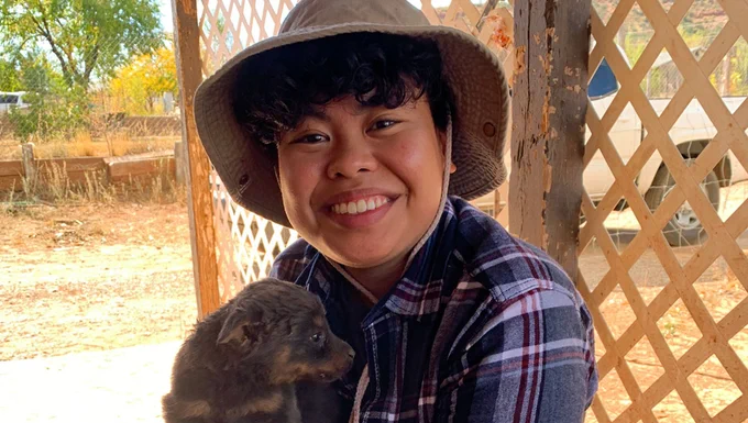 A person wearing a brown hat and a plaid blue shirt sits outside in front of a trellis, holding a brown puppy while smiling.