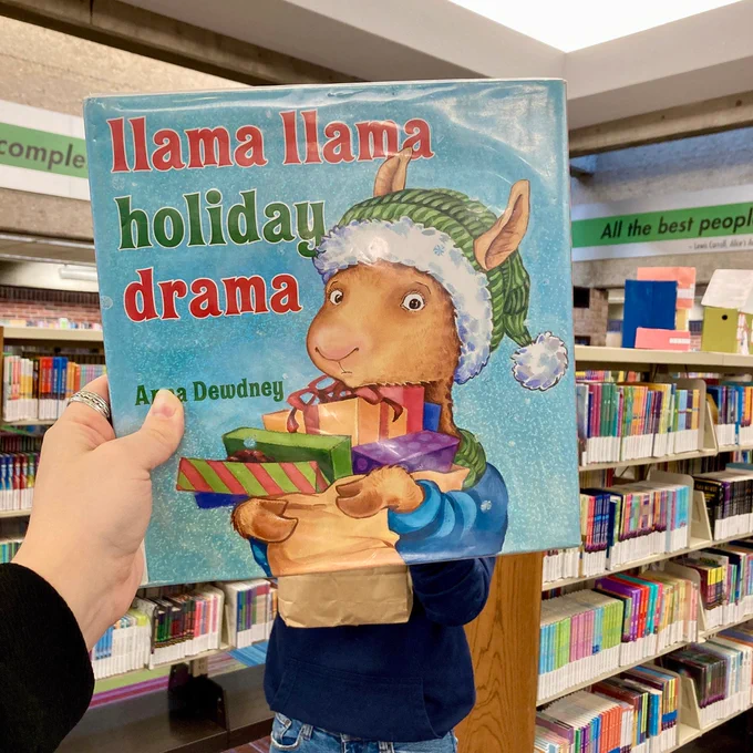 Bookface cover: Llama holding bag of presents in blue coat and green hat standing in book aisle at library