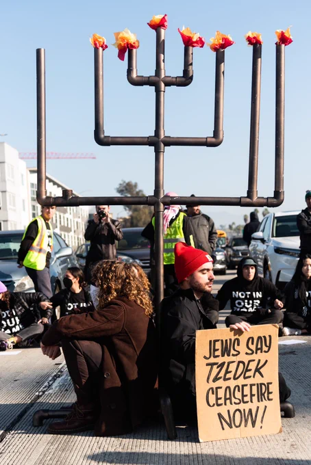 Two people sitting at the base of a very tall menorah in the middle of a freeway. One of them is holding  a sign that says Jews Say Tzedek Ceasefire Now! Behind them is a line of Jews and allies sitting arm in arm in front of cars.