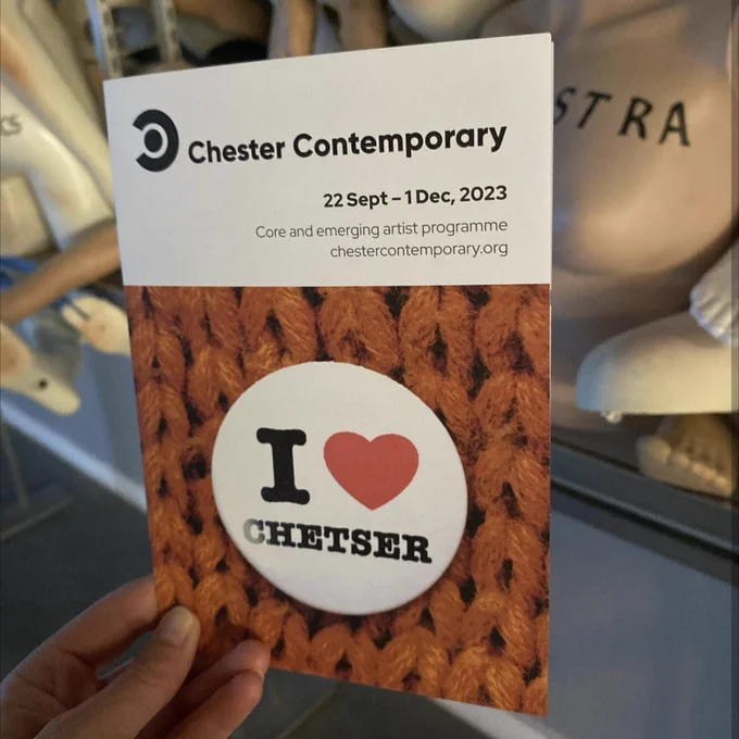 A photo of a hand holding a Chester Contemporary booklet. In the background are the mannequins from the DISARM installation by Fiona Banner aka The Vanity Press.