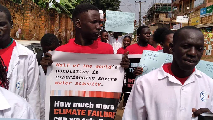 Activists and scientists wearing white lab coats with the extinction symbol coloured in the warming stripes. They are marching along a road in Kampala, Uganda. The placard says: "Half of the world's population is experiencing severe water scarcity."