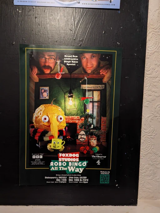 A poster for Foxdog Studios Robo Bingo all the way. Lloyd and Peter loom over a Christmas diaorama featuring a yellow anthropomorphic bingo ball drinking a hot drink by the fire. Lloyd and Pete shaped baubles are on the tree.