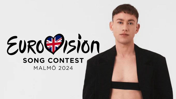 Olly Alexander wears a black jacket with a black strap across the chest and stands beside a Eurovision Song Contest 2024 logo. The v in Eurovision is shaped like a heart and features the red, white and blue Union Jack flag of the United Kingdom.