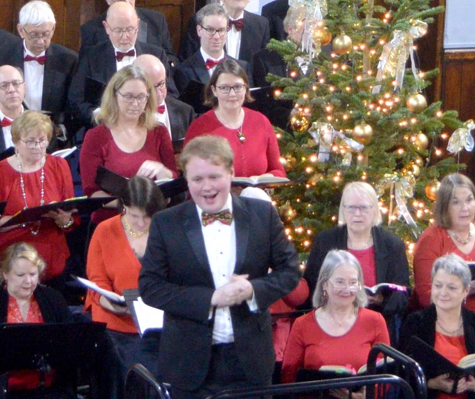 Harry introducing a carol with the choir (in red and black) behind with Christmas tree