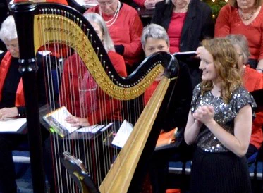 Harpist Alis Huws introducing one of her solos alongside her harp, choir behind