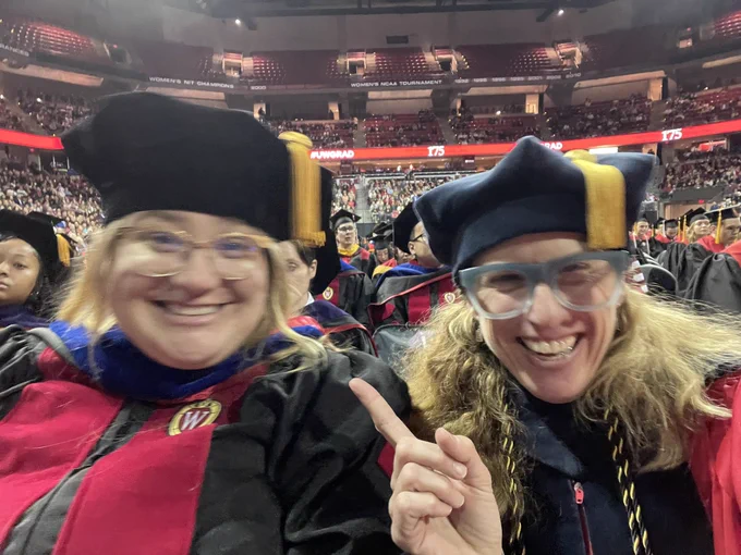 Two blonde ladies in regalia grin their faces off. Kohl Center beyond.