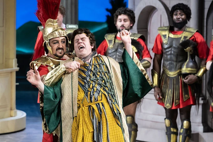 A photo showing a group of performers in Roman clothing, on stage, one holding a vintage knife to the other's throat