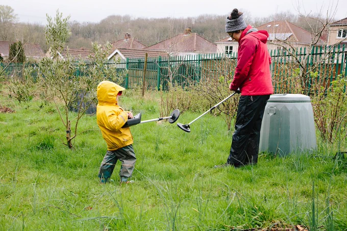 A child in a yellow raincoat and an adult in a red jacket using metal detectors on a grassy field with houses in the background.