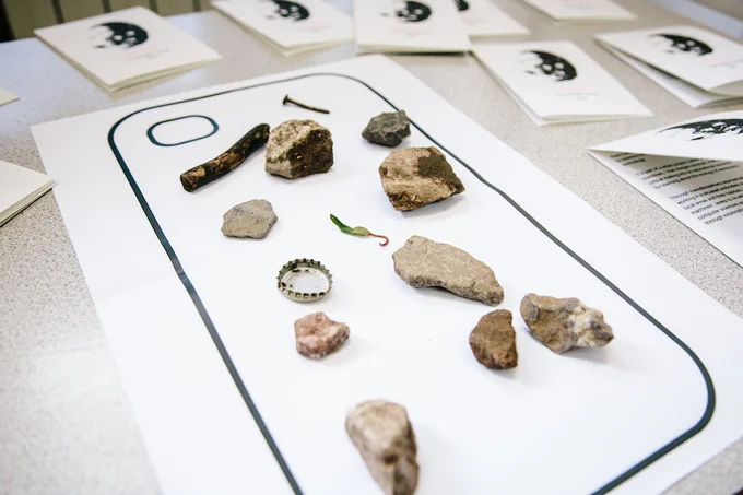 A table with a large paper outline of a mobile phone, scattered with various rocks, a bottle cap, and a screw.